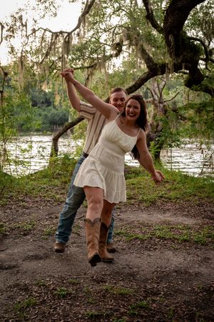 A joyful couple poses outdoors near a river under a moss-covered oak tree, woman in white dress and cowboy boots.