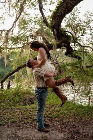 A man lifts a woman in cowboy boots near a mossy oak tree by a serene river in a romantic outdoor setting.