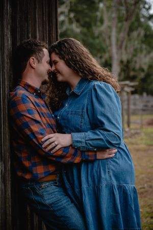 A couple in jeans shares a romantic moment leaning against a rustic wooden barn wall outdoors.