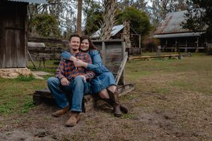 Couple sitting together on rustic farm bench, wearing jeans and flannel, smiling in rural countryside setting.