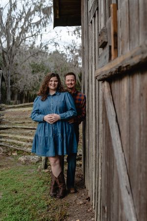 Couple posing by rustic barn in denim dress and plaid shirt, surrounded by moss-draped trees in a countryside setting.