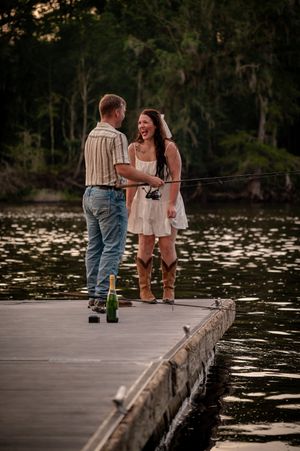 Couple laughing on a dock by a lake, holding a fishing rod with champagne bottles nearby at sunset.