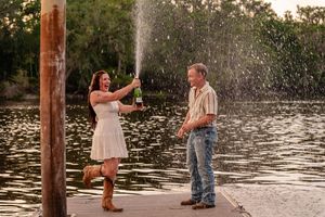 Couple celebrating on a dock with champagne spray over a lake at sunset, woman in white dress and boots.