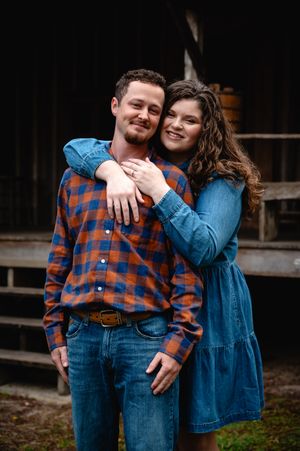 A smiling couple poses outdoors, woman in blue dress hugging man in plaid shirt and jeans near a rustic barn.