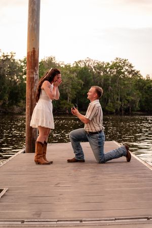 Man proposes on a wooden dock at sunset, kneeling with ring box as woman covers her mouth in surprise.