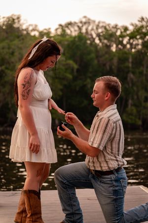 Man kneeling on a dock proposing to a woman in a white dress and cowboy boots near a lake at sunset.