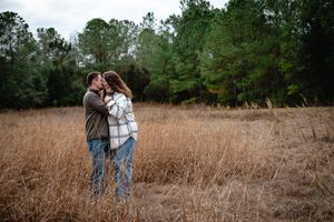 Couple sharing a romantic kiss in a golden field surrounded by lush green trees during an outdoor engagement session.
