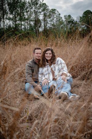Couple sitting together in tall golden grass field, wearing casual fall outfits, smiling, with pine trees in background.