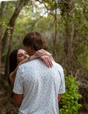 A couple embraces in a lush forest setting, the woman smiling as she wraps her arms around the man.
