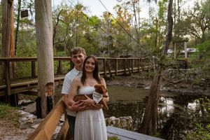 Couple embracing on a wooden bridge surrounded by lush trees and a serene creek in a romantic outdoor setting.