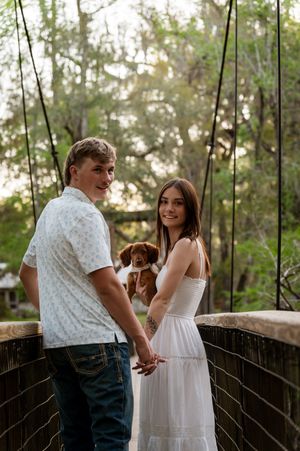 A couple holding hands on a bridge with a brown puppy, surrounded by lush green trees.
