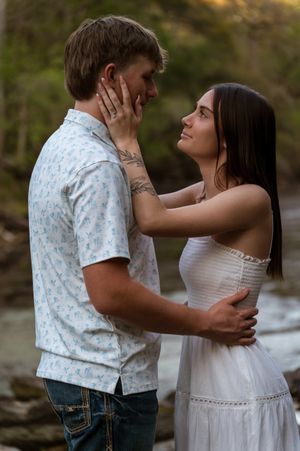A young couple shares a romantic moment outdoors, with the woman gently touching the mans face while smiling up at him.
