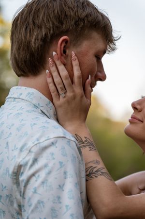 A woman with a tattoo and engagement ring tenderly holds a man's face during a romantic outdoor couples photo session.