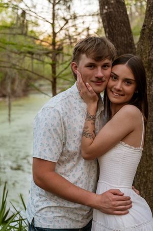 A young couple poses romantically near a tree by a pond, the woman in white dress embracing the man in a floral shirt.