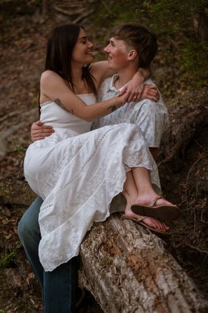 Couple sitting on a fallen log in the woods, embracing and smiling at each other, woman in white dress.