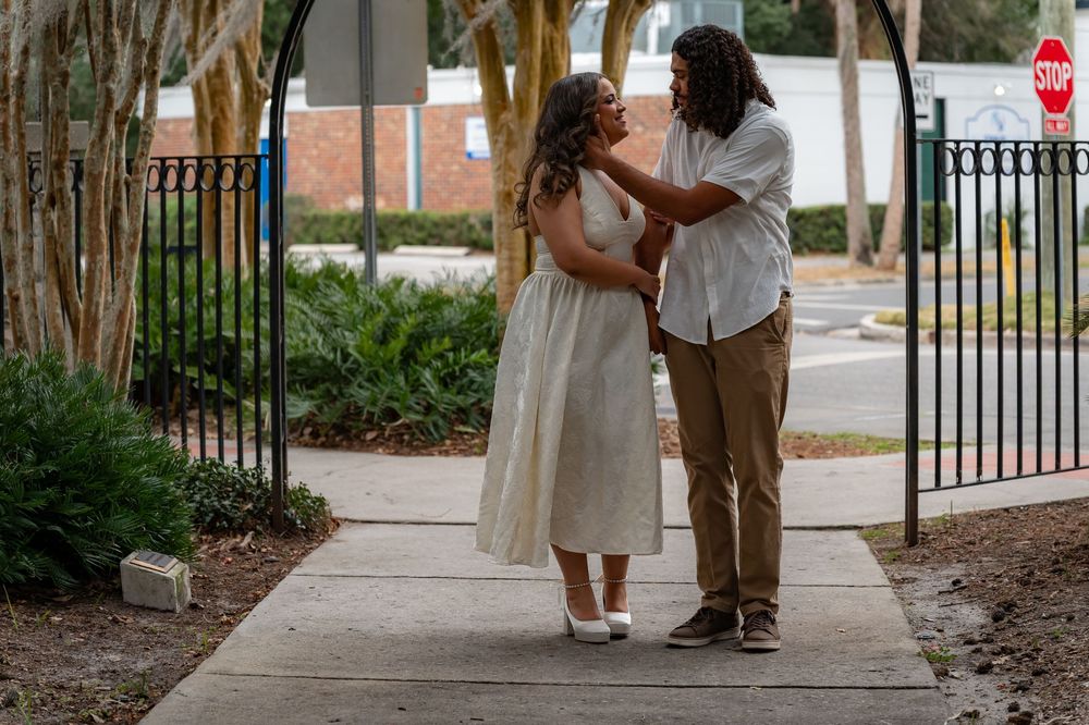 Couple sharing a romantic moment under a garden arch on a sunlit pathway, woman in white dress, man in casual attire.
