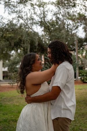 A couple embraces outdoors near trees, woman in white dress gazing up at curly-haired man in white shirt.