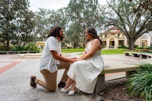 Man kneeling on one knee proposing to woman seated on bench in scenic park setting.