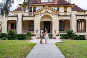 Couple embracing on a walkway in front of an elegant historic Southern mansion with lush green landscaping.