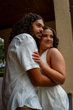 A couple in white outfits embrace tenderly near a column, the man with curly hair holding the smiling woman close.