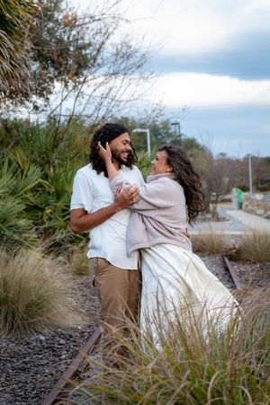 A joyful couple embraces and smiles at each other near rustic train tracks surrounded by wild grasses under a cloudy sky.