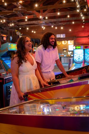 A joyful couple in white outfits plays pinball at a vintage arcade with warm string lights overhead.