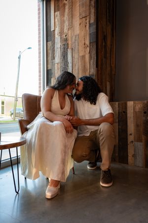 Couple sharing an intimate moment, touching foreheads while holding hands near a window with rustic wood wall backdrop.