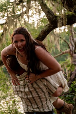 Two women laughing and playing piggyback in a lush forest setting with moss-draped trees.