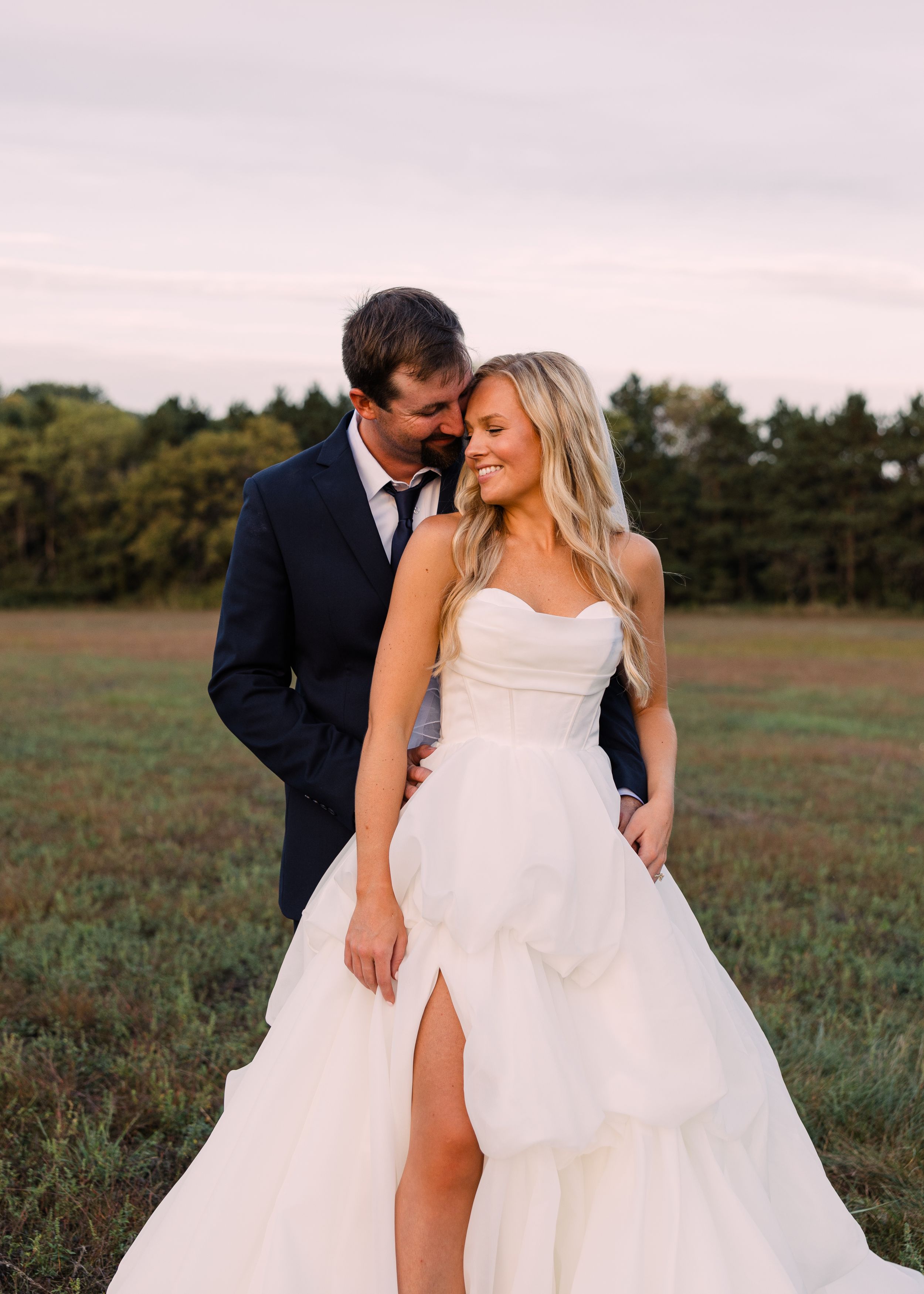 Groom in a navy suit embraces a smiling bride in a strapless white gown with a high slit in a grassy field at sunset.