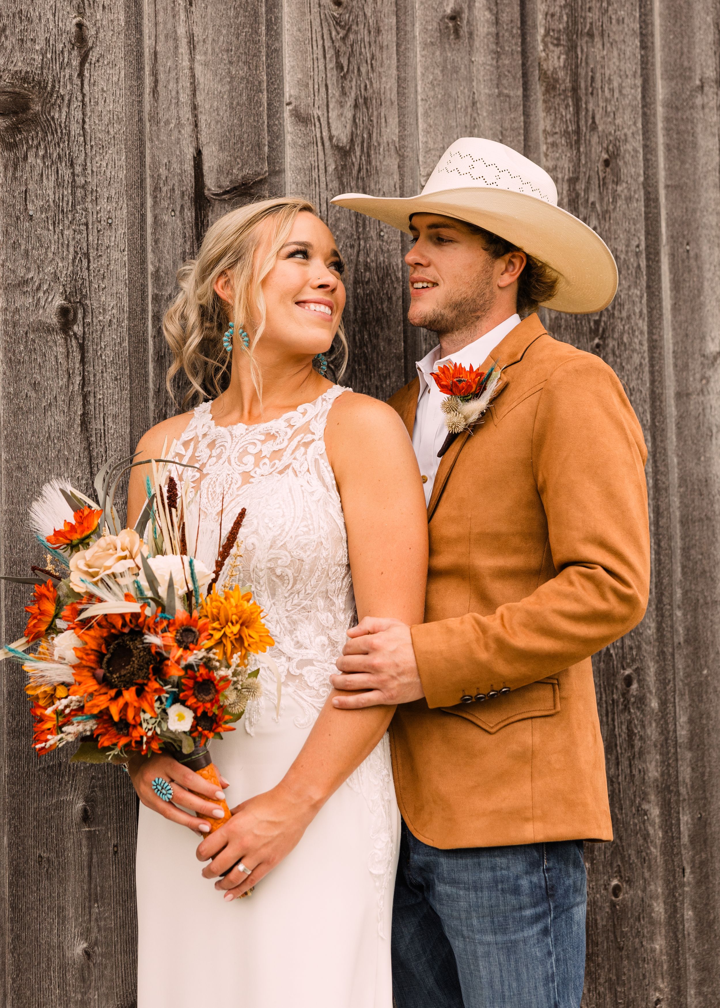 Smiling bride holding an orange sunflower bouquet embraced by a groom in a cream cowboy hat and tan jacket by a rustic barn wall.