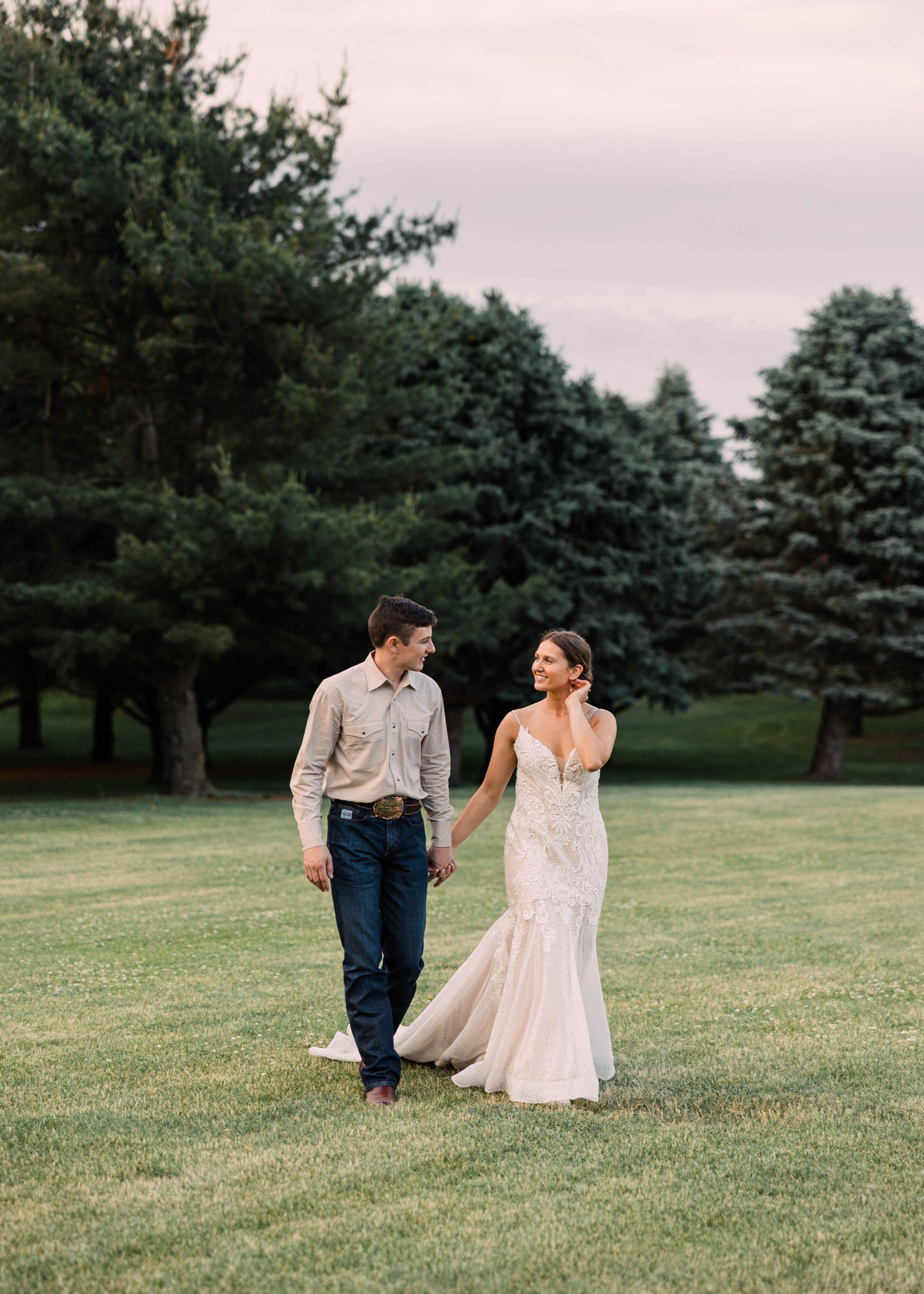 Bride in a lace wedding gown walks hand in hand with groom in a button-up shirt across a lawn lined with evergreen trees.