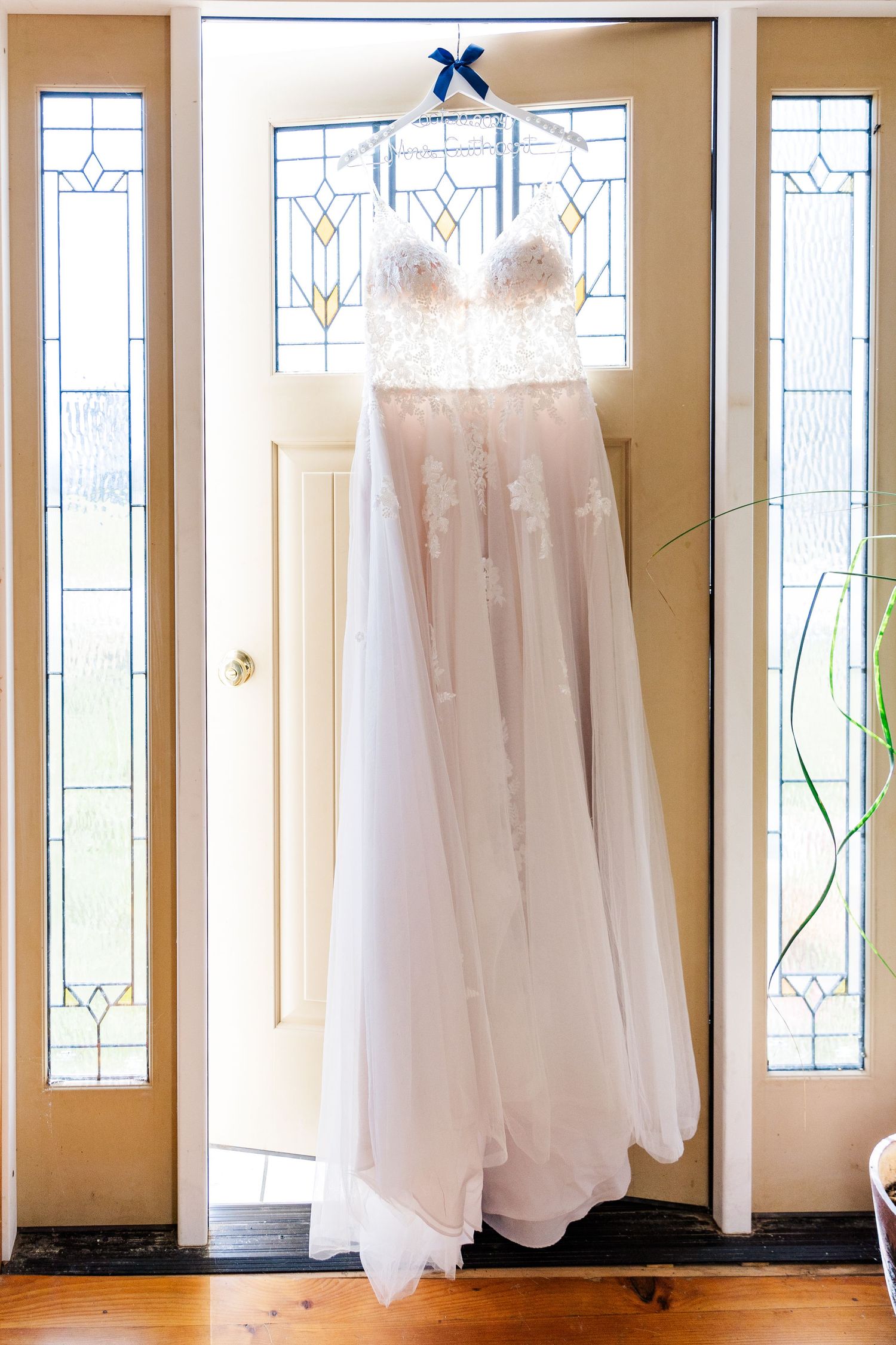 Elegant wedding gown hanging in front of traditional stained glass windows with blue bow detail.