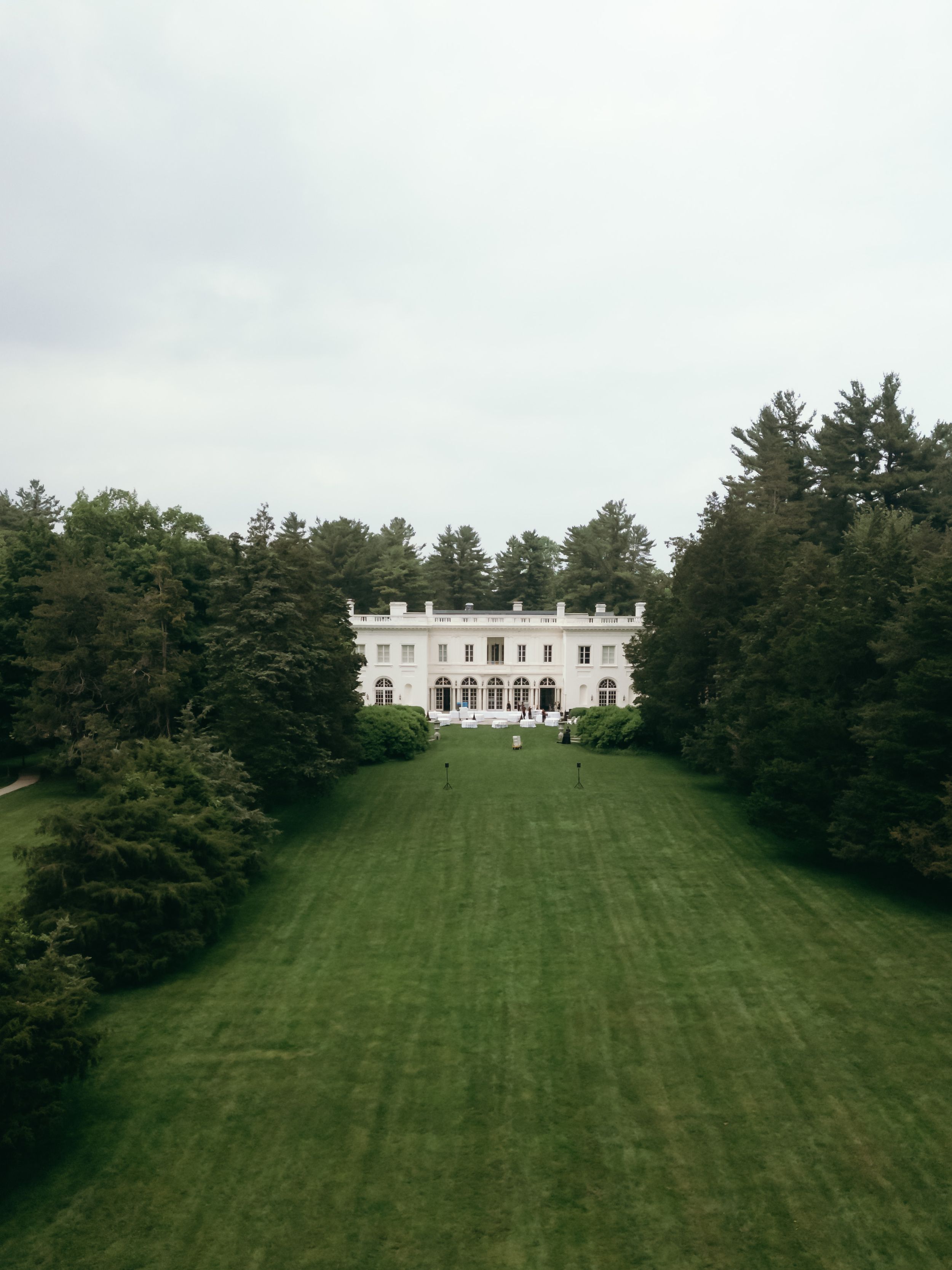 A grand white mansion sits atop a manicured green hillside surrounded by tall evergreen trees.