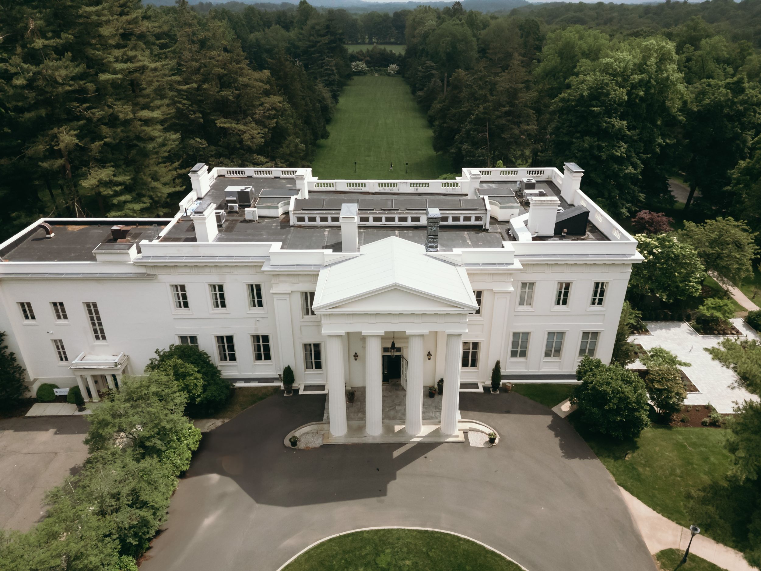 Aerial view of a grand white neoclassical mansion with symmetrical wings and manicured gardens surrounded by trees.