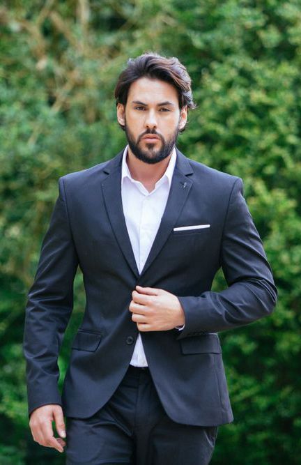 A man in a black suit and white shirt poses outdoors against a green natural background.
