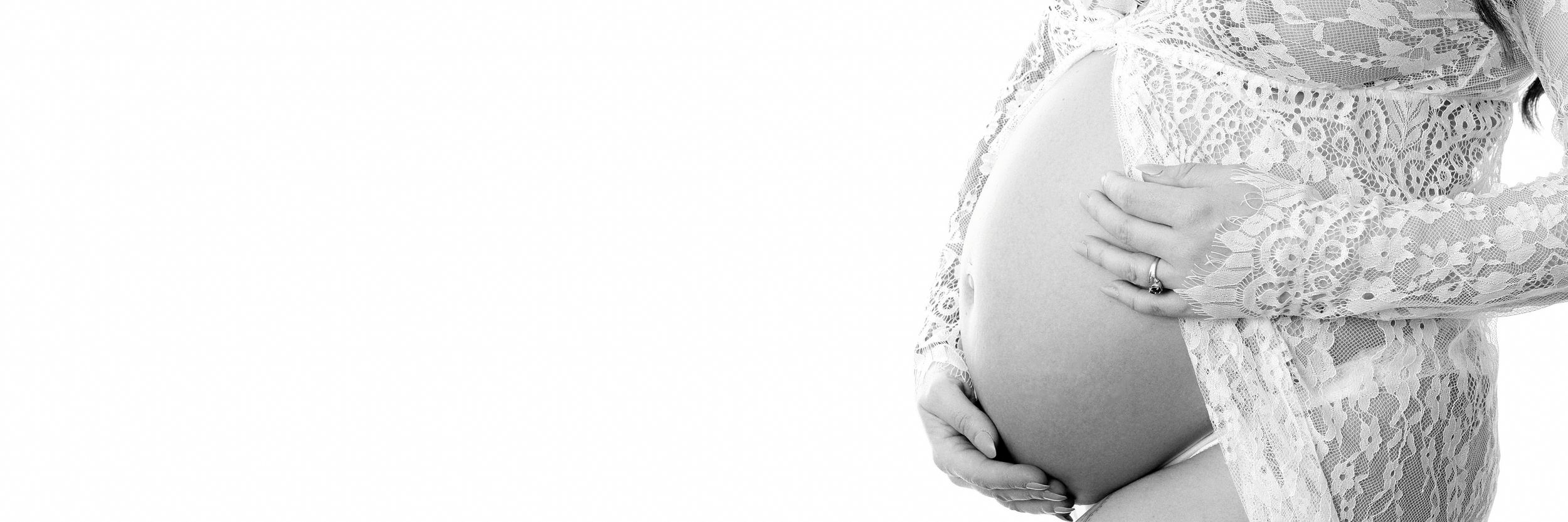 A pregnant photoshoot showing a woman wearing a white patterned lace gown. She is cupping her hands around her baby like a mother would.