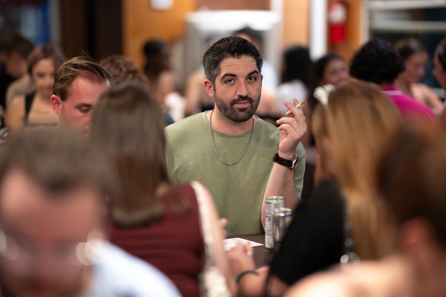Man in green t-shirt gesturing while talking at a crowded indoor dining event with guests in background.