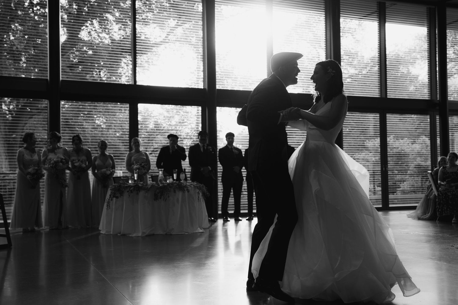 Silhouettes of wedding guests and dancers cast dramatic shadows in a large reception venue with floor-to-ceiling windows.