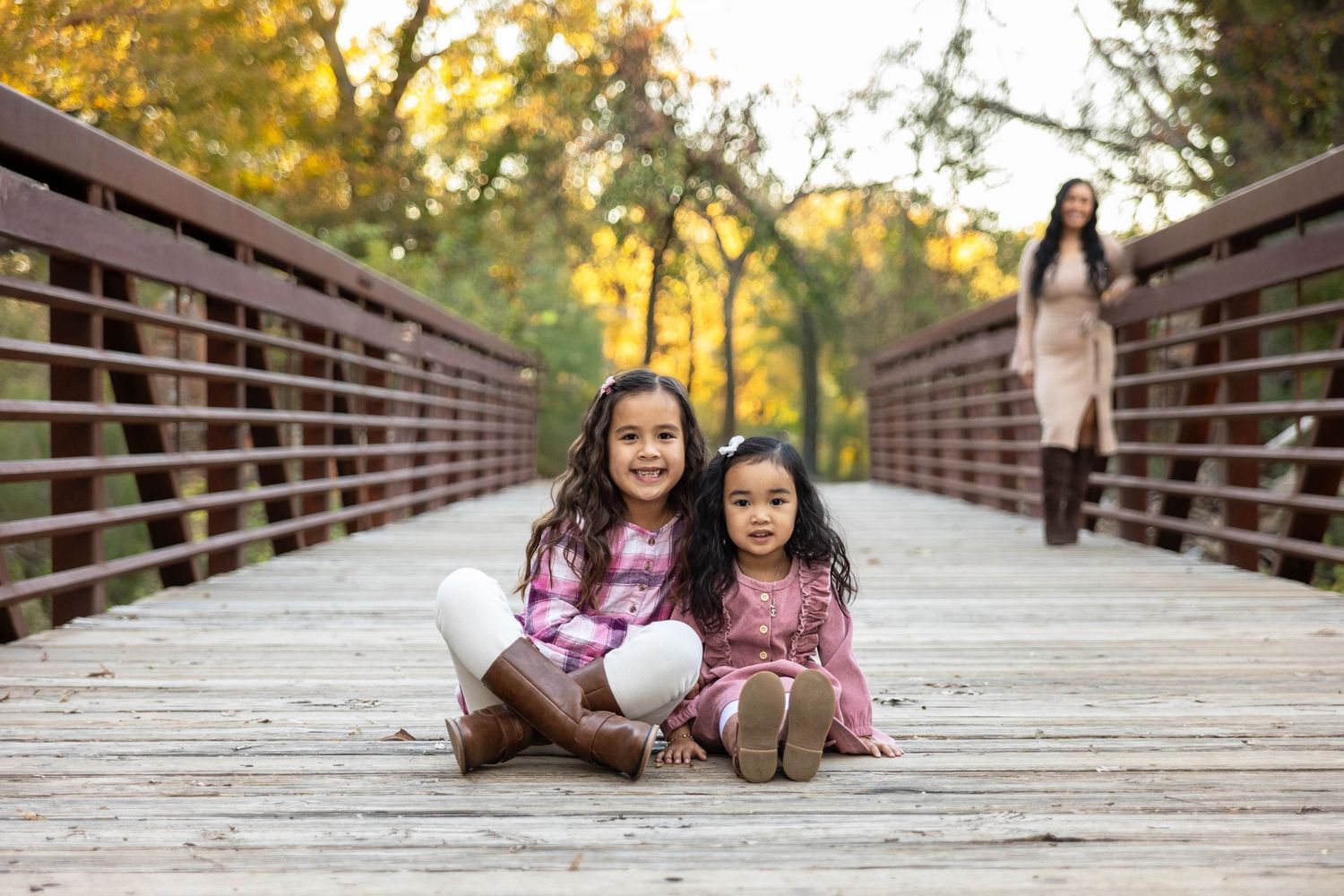 Two young girls sitting on a bridge with their Mother watching in the background.