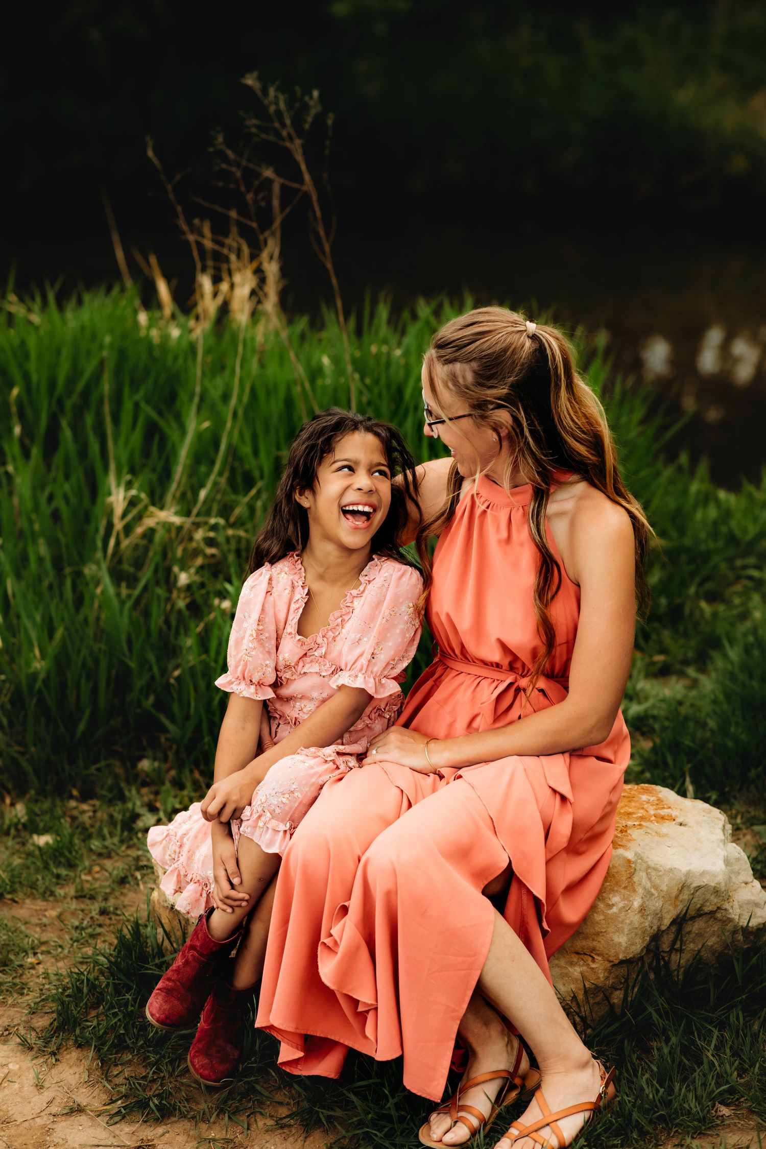 Two women in coral and pink dresses share joyful laughter while sitting together on rocks in a grassy outdoor setting.