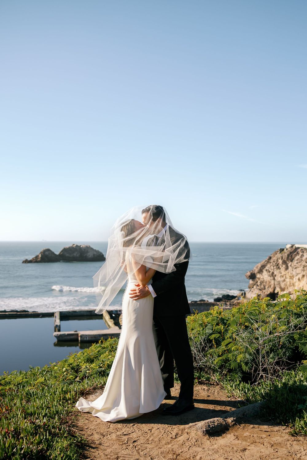 A couple kisses on a scenic coastal cliff overlooking the ocean during their wedding portraits as the veil flows in the wind at Sutro Baths.