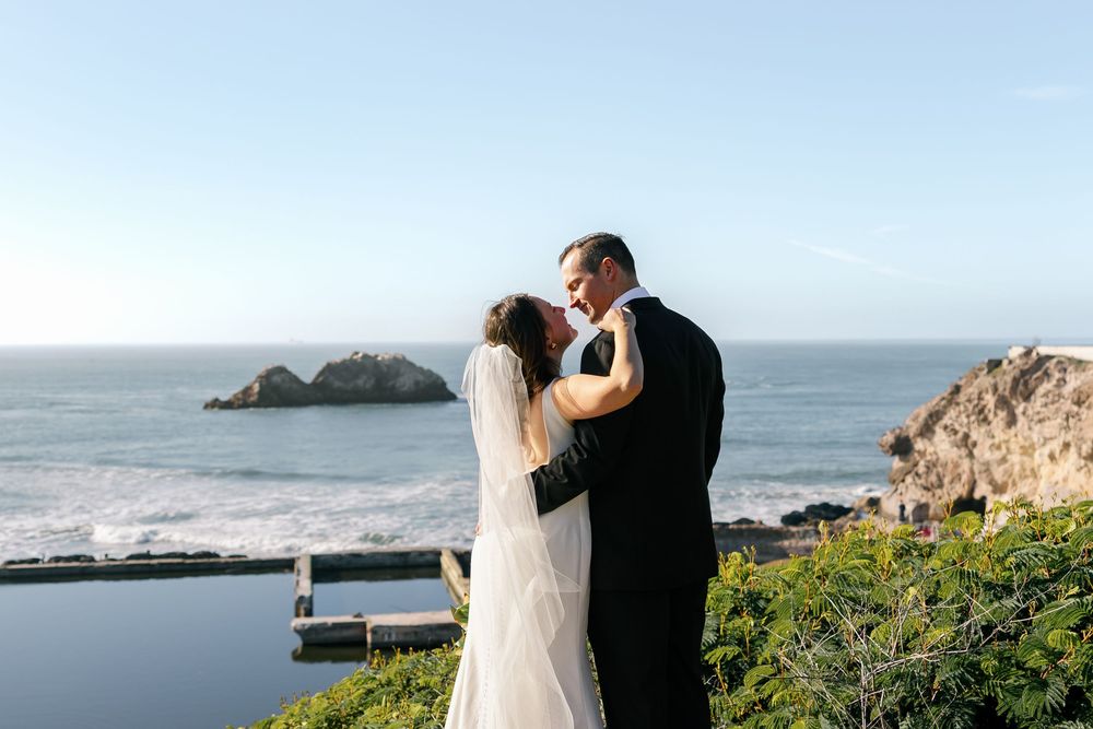 A couple shares a romantic moment on a scenic coastal cliff overlooking the ocean during their wedding photoshoot.