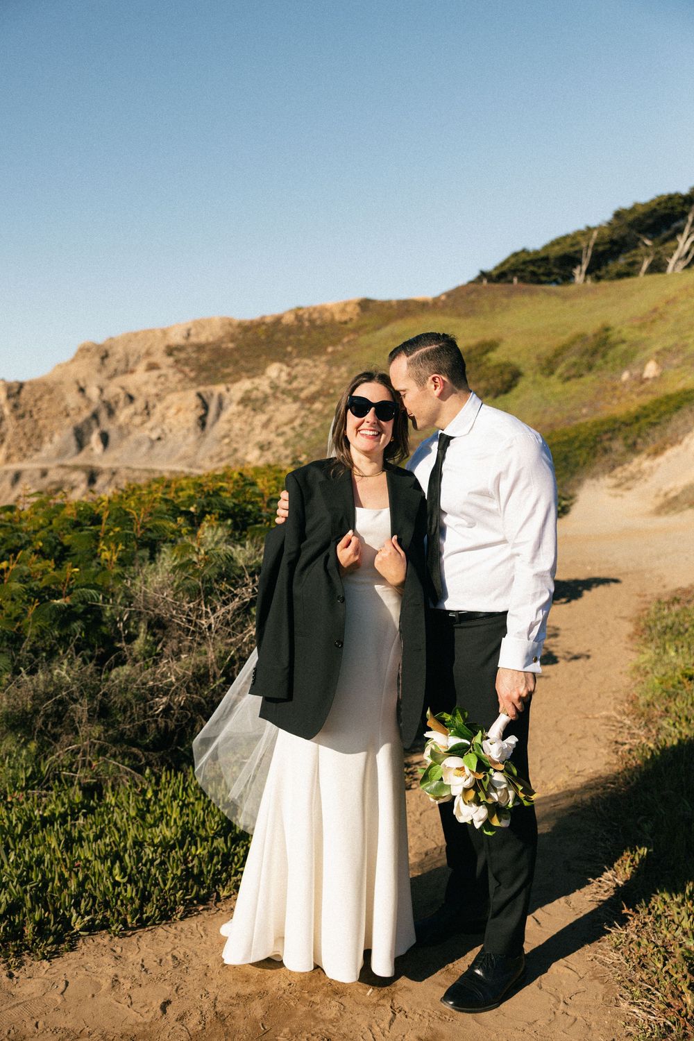 A couple shares a fun moment on a coastal trail with cliffs and ocean views, holding a bouquet of white flowers and wearing sunglasses.