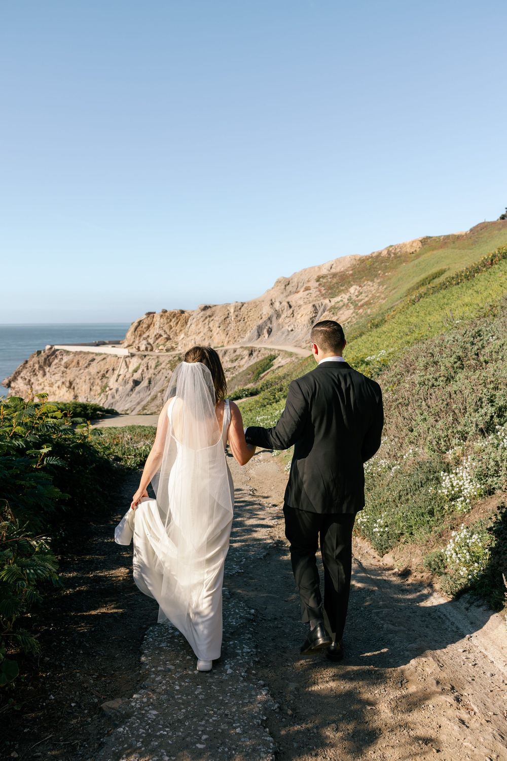 A couple in wedding attire walking along a scenic coastal trail with dramatic cliffs and ocean views.