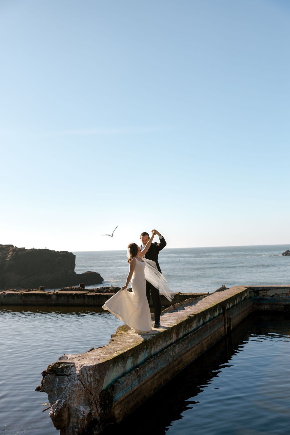 A couple stands on a concrete pier overlooking the ocean in full sun in a romantic coastal setting at Sutro Baths.