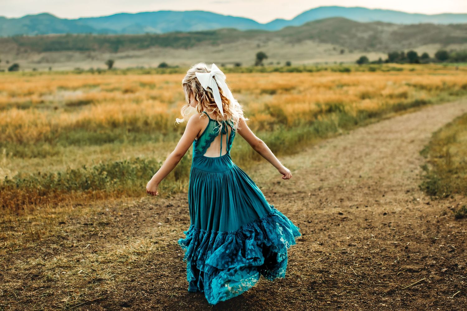 A figure in a flowing teal dress walks along a dirt path through golden grasslands with mountains in the distance.