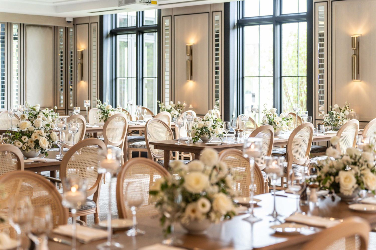 Formal dining room with tall windows and elegant table settings featuring white floral centerpieces.