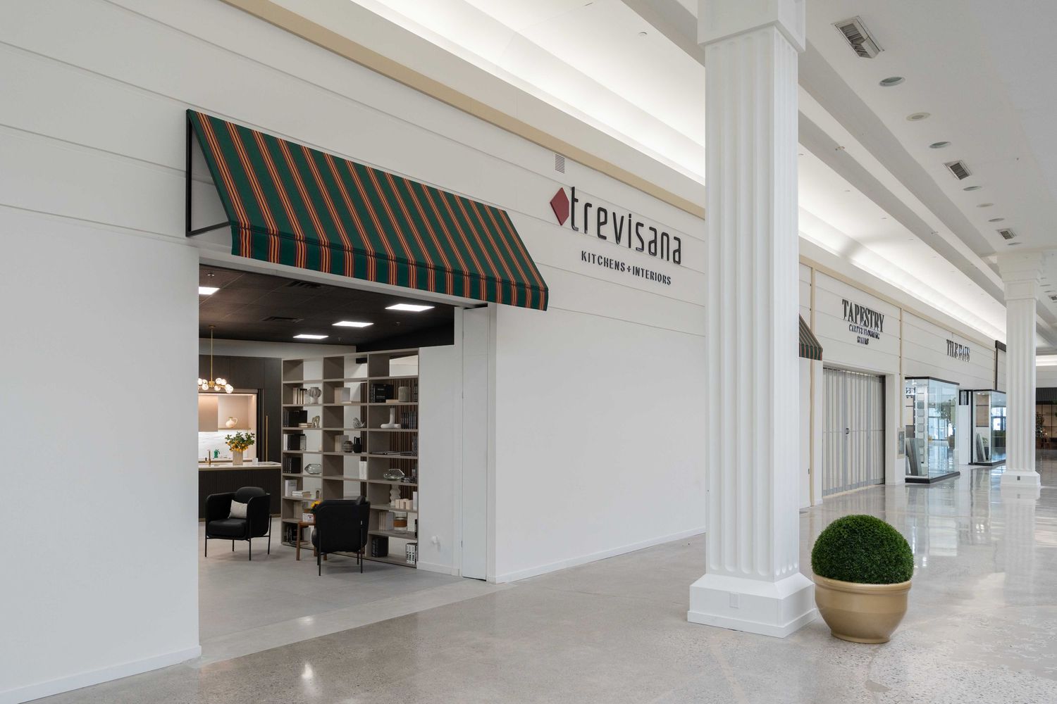 Modern shopping mall interior with white walls, columns and decorative potted plants along a bright retail corridor.