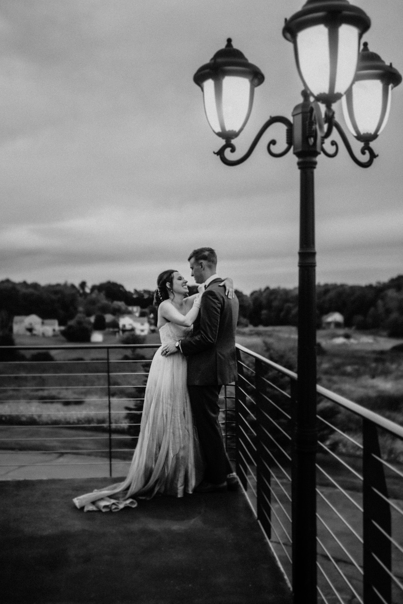 A romantic black and white wedding photo of a couple embracing on a balcony with vintage lamp posts at dusk.