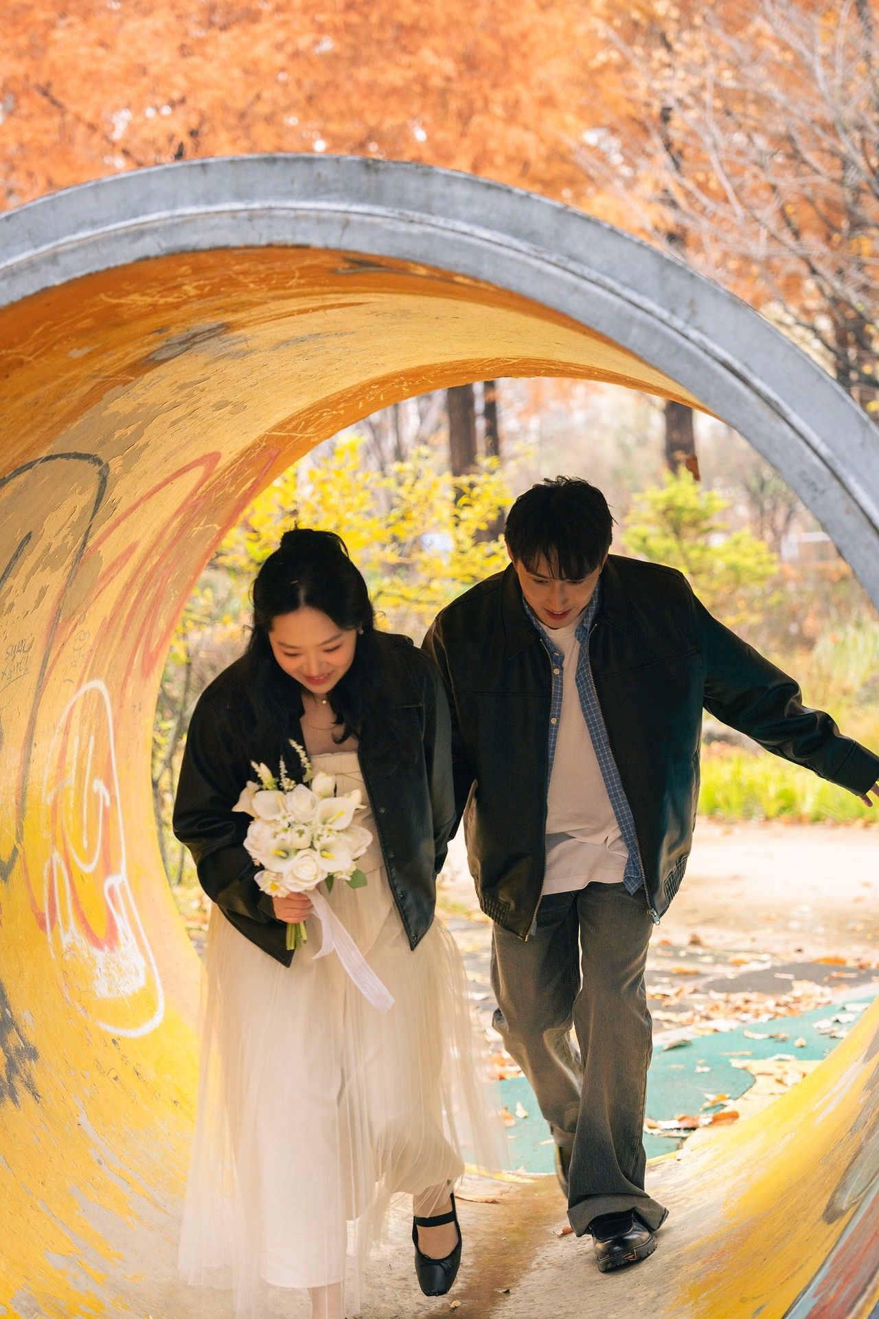 A couple in wedding attire poses playfully inside a yellow-lit tunnel surrounded by autumn leaves.